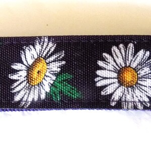 May include: A daisy-patterned key fob with a gold-colored metal clasp and key ring. The fabric features white daisies with yellow centers and green leaves against a dark blue background. The key fob is a practical and decorative accessory.