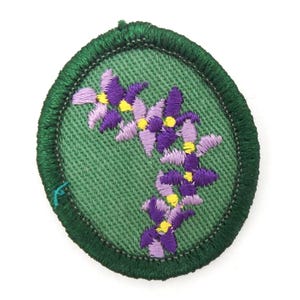 May include: A circular embroidered patch featuring a light green fabric background with a dark green stitched border. The center displays a cluster of purple and light purple flowers with small yellow centers arranged in a curved, diagonal pattern.