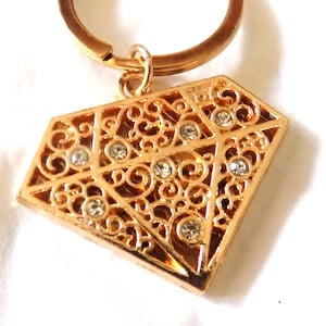 May include: Gold-colored diamond-shaped keychain pendant with a filigree design and small, clear rhinestones. The pendant is attached to a gold-colored key ring. The design features a geometric pattern with swirls and clear crystals.