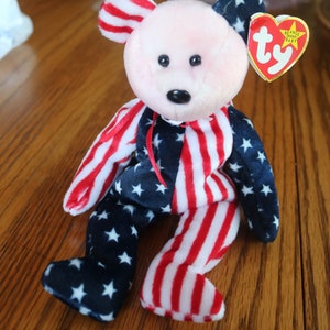 May include: A red, white, and blue beanie baby bear wearing a red, white, and blue striped outfit with white stars. The bear has a pink nose and black eyes. The bear is sitting on a wooden surface. The bear has a Ty tag attached to its ear that says "Beanie Babies Baby".