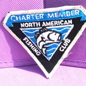 May include: A triangular patch with white stitching around the edges. The patch is black with a blue and white fish graphic in the center. The text "Charter Member North American Fishing Club" is embroidered in white on the patch.