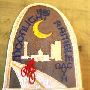 May include: A gray embroidered patch with white stitching. The patch features a crescent moon, a cityscape, a cyclist, and the text "Moonlight Ramble 1980".