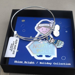 May include: Silver bangle bracelet with a charm featuring a cartoon elf holding a sign that says "North Pole". The bracelet is in a black box with a blue and white card that says "Alex and Ani" and "Shine Bright / Holiday Collection".