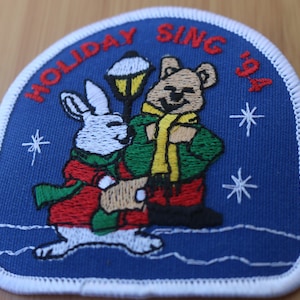 May include: A blue embroidered patch with a white border. The patch features a white rabbit, a brown bear, and a street lamp. The text "HOLIDAY SING 94" is embroidered in red on the patch.