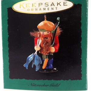 May include: A Keepsake Ornament collector's series box featuring a fisherman nutcracker. The nutcracker is wearing a red coat, green hat, and blue boots, holding a blue fish and a fishing rod. The box has a green border and the text "Keepsake Ornament".
