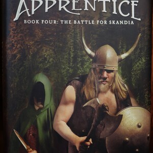 May include: Book cover for "Ranger's Apprentice: Book Four: The Battle for Skandia" by John Flanagan. The cover features a Viking warrior with a horned helmet, holding an axe and shield, and a hooded figure in the background.
