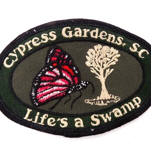 May include: An oval embroidered patch with an olive green center and a black border. It features a red and black butterfly, a beige tree, and the text "Cypress Gardens, SC" at the top and "Life's a Swamp" at the bottom in beige embroidery.