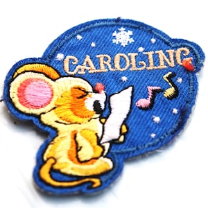 May include: A blue felt patch with a yellow mouse singing carols. The mouse is holding a sheet of music and there are musical notes in the background. The text "CAROLING" is embroidered in white on the patch.