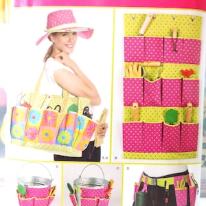 May include: Simplicity sewing pattern featuring a tote bag, bucket, and apron with pockets, all in a floral and polka dot design. The pattern includes a sun hat with a pink polka dot pattern. The text "Simplicity" is visible.