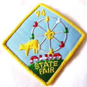 May include: Embroidered patch with a light blue background and a yellow border. The patch features a Ferris wheel, a yellow cow, and the words "STATE FAIR" in green. The number "94" is at the top.
