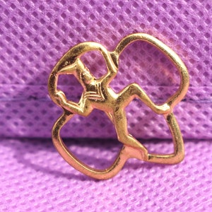 May include: A gold-colored pin featuring a stylized figure in a dynamic pose, set within a decorative, openwork design. The pin has a shiny finish and is set against a purple textured background.