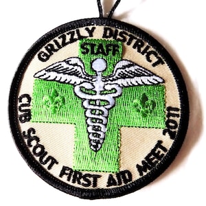 May include: Round embroidered patch with the text "GRIZZLY DISTRICT CUB SCOUT FIRST AID MEET 2011". The patch features a green cross with a white medical symbol and wings. The patch is tan with a black border.