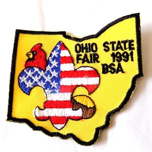 May include: Embroidered patch in the shape of Ohio with a yellow background and black border. The patch features a fleur-de-lis design with red, white, and blue elements, a cardinal, and text that reads "OHIO STATE FAIR 1991 BSA."