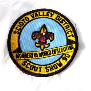 May include: A circular embroidered patch with a light blue center and a yellow border. It features a brown and gold fleur-de-lis emblem with a red, white, and blue shield. Text reads: SCIOTO VALLEY DISTRICT, WONDERFUL WORLD OF SCOUTING, SCOUT SHOW 90.