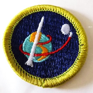May include: Embroidered patch with a blue background and a yellow border. The patch features a rocket, a planet, and a satellite, with red and orange accents. The design is circular and has a detailed, stitched appearance.