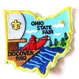 May include: Embroidered patch in the shape of Ohio, featuring a red cardinal bird, the text "OHIO STATE FAIR", and "DISCOVER 1992". The patch has a yellow border and includes a fleur-de-lis symbol.