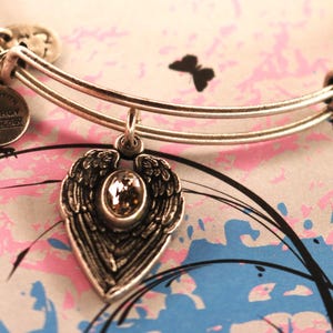 May include: Silver charm bracelet with a heart-shaped charm featuring a pair of angel wings and a clear crystal. The charm is attached to the bracelet with a silver loop. The bracelet is inscribed with "Inspired with Cleveroy Technology".