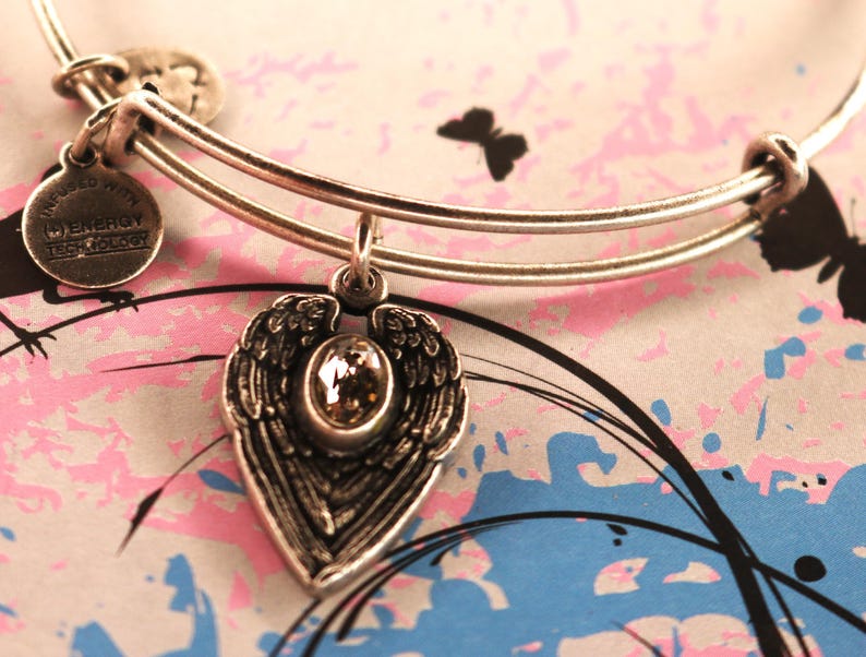 May include: Silver charm bracelet with a heart-shaped charm featuring a pair of angel wings and a clear crystal. The charm is attached to the bracelet with a silver loop. The bracelet is inscribed with "Inspired with Cleveroy Technology".
