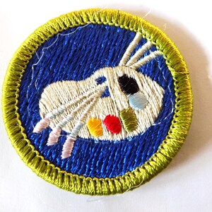 May include: Embroidered patch featuring a white design on a blue background, with a yellow border. The design includes a stylized image with colorful accents of yellow, red, black, and blue. The patch is round and likely for clothing or accessories.