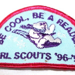 May include: A Girl Scouts patch with a light blue background and a red border. The patch features a cartoon bear wearing a scarf and sledding. Text on the patch reads "BE COOL, BE A READER" and "GIRL SCOUTS '96-'97".