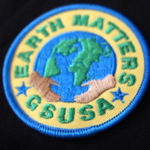 May include: A yellow and blue embroidered patch with the text "Earth Matters GSUSA" and a depiction of a globe with two hands holding it.