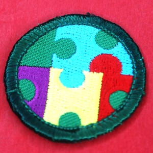 Pode incluir: Um remendo redondo com borda verde com um design de pe&ccedil;as de quebra-cabe&ccedil;a coloridas. As pe&ccedil;as do quebra-cabe&ccedil;a s&atilde;o vermelhas, amarelas, azuis, verdes e roxas. Cada pe&ccedil;a tem um padr&atilde;o de pequenos c&iacute;rculos verdes.