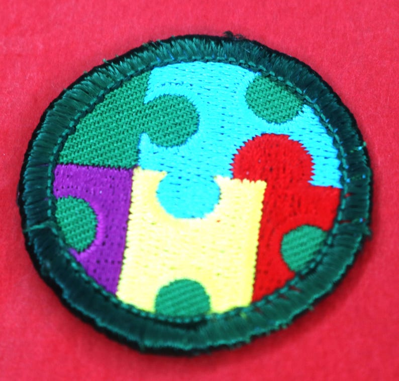 Pode incluir: Um remendo redondo com borda verde com um design de pe&ccedil;as de quebra-cabe&ccedil;a coloridas. As pe&ccedil;as do quebra-cabe&ccedil;a s&atilde;o vermelhas, amarelas, azuis, verdes e roxas. Cada pe&ccedil;a tem um padr&atilde;o de pequenos c&iacute;rculos verdes.