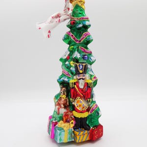 May include: A Christmas tree ornament featuring a green tree shape with a red and gold nutcracker soldier and a small dog. The ornament is decorated with colorful presents and a white ribbon.
