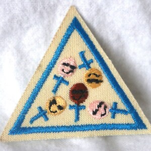 May include: A triangular Girl Scout patch with a blue border and a white background. The patch features five embroidered smiling faces in different colors, surrounded by blue crosses.