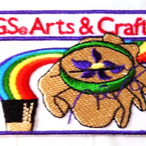 May include: Embroidered patch with the text "GS Arts & Crafts" in red. The patch features a rainbow, a hand holding a flower, and a black container. The patch has a white background with a purple border.