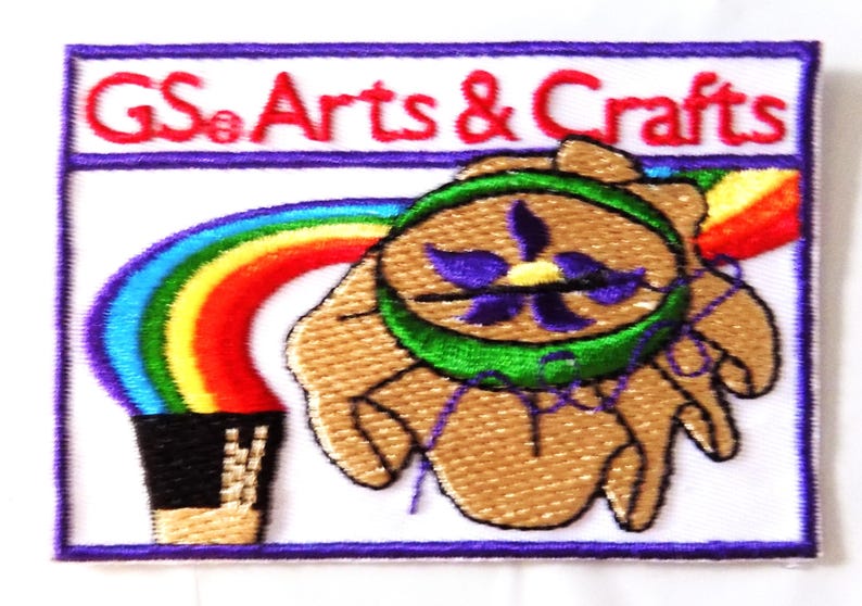 May include: Embroidered patch with the text "GS Arts & Crafts" in red. The patch features a rainbow, a hand holding a flower, and a black container. The patch has a white background with a purple border.
