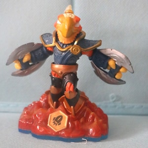 May include: A gold, blue, and red Skylanders figure of a bird-like creature with wings. The figure is standing on a red and orange base with a rocket symbol.