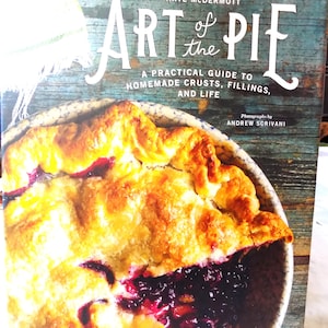 May include: A cookbook titled "Art of the Pie" by Kate McDermott. The cover features a close-up of a golden-brown pie with a dark berry filling. The book's subtitle reads "A Practical Guide to Homemade Crusts, Fillings, and Life."