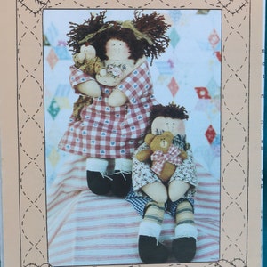 May include: A sewing pattern for two rag dolls, Nellie and Ned, standing together. The dolls are dressed in gingham and floral fabric. The pattern is titled "Homespun at Heart Designs" and includes the number "HS 13". The dolls are 9 1/2 inches tall.
