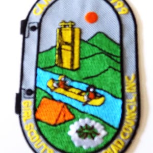 May include: Embroidered patch with a camping theme. The patch features a yellow raft with two people, a yellow suitcase, an orange tent, and a campfire. Text includes "CAMP ROCKS! 1999" and "GIRL SCOUTS - TRIAD COUNCIL INC."