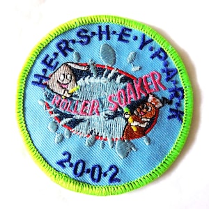 May include: A circular embroidered patch with a light blue background and a bright lime green border. It features the text HERSHEYPARK, ROLLER SOAKER, and 2002. The design includes two cartoon characters in a water splash graphic with blue details.