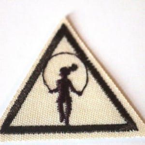 May include: A triangular patch with a cream-colored background and a black border. The patch features a silhouette of a person jumping rope inside a circle. The design is embroidered, suggesting a fabric patch.