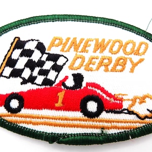 May include: Embroidered patch with a green border and a white background. The patch features a red race car with the number 1, a checkered flag, and the words "PINEWOOD DERBY" in orange thread. The car has black wheels and a trail of smoke.