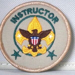 May include: A tan embroidered patch with a green circle around a yellow and brown scout emblem. The text "INSTRUCTOR" is in green around the circle.