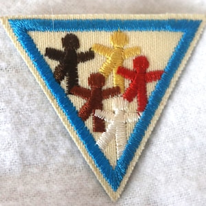 May include: A triangular patch with a blue border and five stick figures of different colors holding hands. The figures are brown, yellow, red, brown, and white.