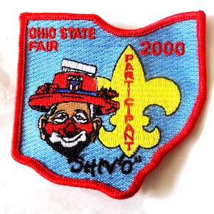 May include: Embroidered patch in the shape of Ohio with a red border. The patch features a clown with a red hat, the text "OHIO STATE FAIR 2000", a yellow fleur-de-lis, and the word "PARTICIPANT".