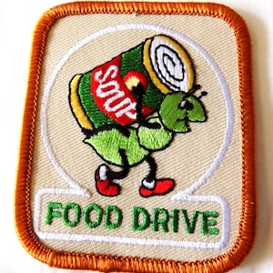 May include: Embroidered patch with a tan background and an orange border. The patch features a green cartoon ant carrying a can of soup. The can is red and green with the word "SOUP" in white. The words "FOOD DRIVE" are in green.