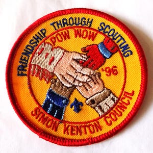 May include: A round, embroidered patch with a yellow background and a red border. The patch features two hands shaking, with text that reads "Friendship Through Scouting Pow Wow '96 Simon Kenton Council."