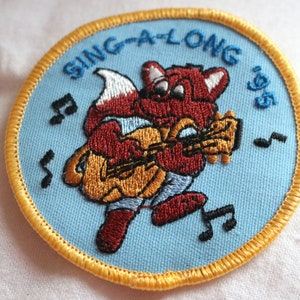 May include: A blue and yellow embroidered patch with a red fox playing a guitar. The text "SING-A-LONG '95" is embroidered around the edge of the patch.