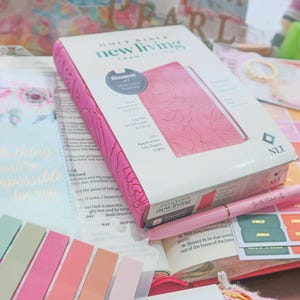 May include: A pink and white Holy Sister New Living Translation Bible with a floral design on the cover. The Bible is open to a page with text and is surrounded by various stationery items, including stickers, bookmarks, and a pen.