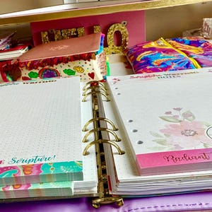 Könnte beinhalten: Offener Planer mit linierten Seiten und floralen Akzenten. Die Seiten tragen die Aufschrift "Girls Write Scripture!" und "Radiant Pearl". Weitere Schreibwaren, darunter Notizbücher und eine Reißverschlusstasche, sind im Hintergrund zu sehen.