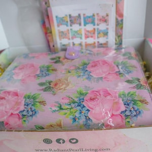 May include: A gift box containing a floral-patterned planner, stickers, and decorative filler. The planner features a pink background with large pink roses and blue hydrangeas. The box is white with a floral design.
