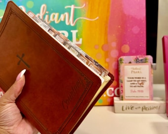 May include: A brown leather-bound book with a cross emblem and labeled tabs. The book is held open, revealing the pages. The background includes colorful artwork and decorative items, suggesting a theme of faith and inspiration.