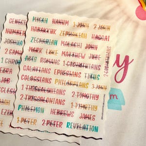 May include: Two stacks of Bible verse labels with colorful text. The labels are arranged in a wavy pattern, with each label displaying a different book of the Bible. The background is a light-colored fabric with colorful accents.