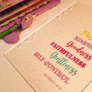 Può includere: Un planner bianco con glitter con le parole "Amore, Gioia, Pace, Pazienza, Gentilezza, Bontà, Fedeltà, Dolcezza, Autocontrollo" scritte in lettere colorate. Il testo "Galati 5:22-23" è scritto in fondo al planner.